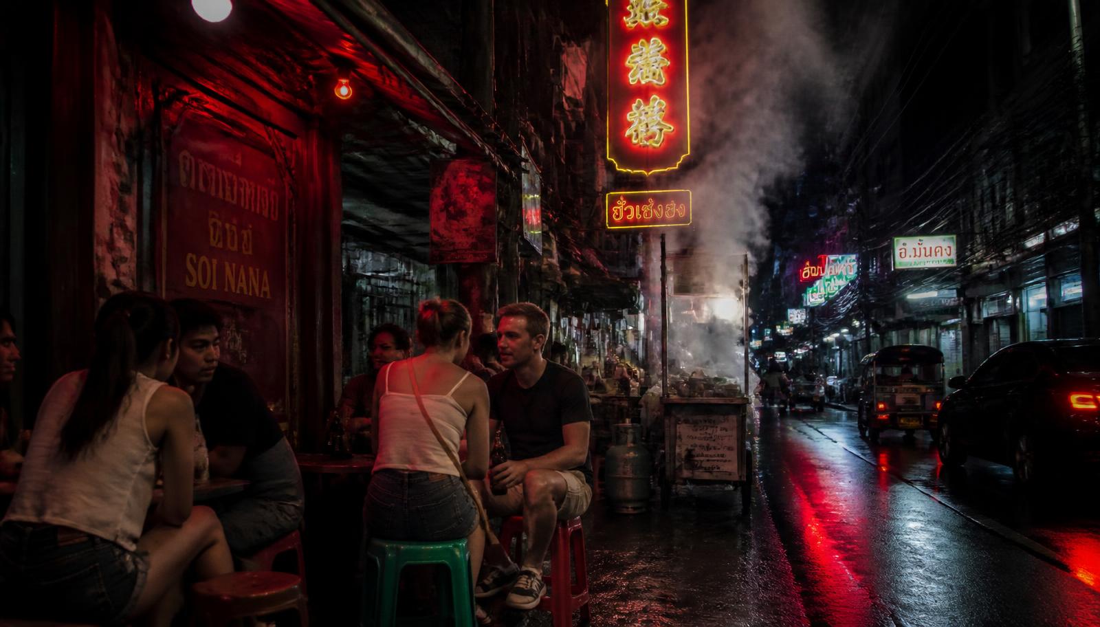 Locals and travellers drinking on plastic stools on a neon-lit Chinatown street in Bangkok